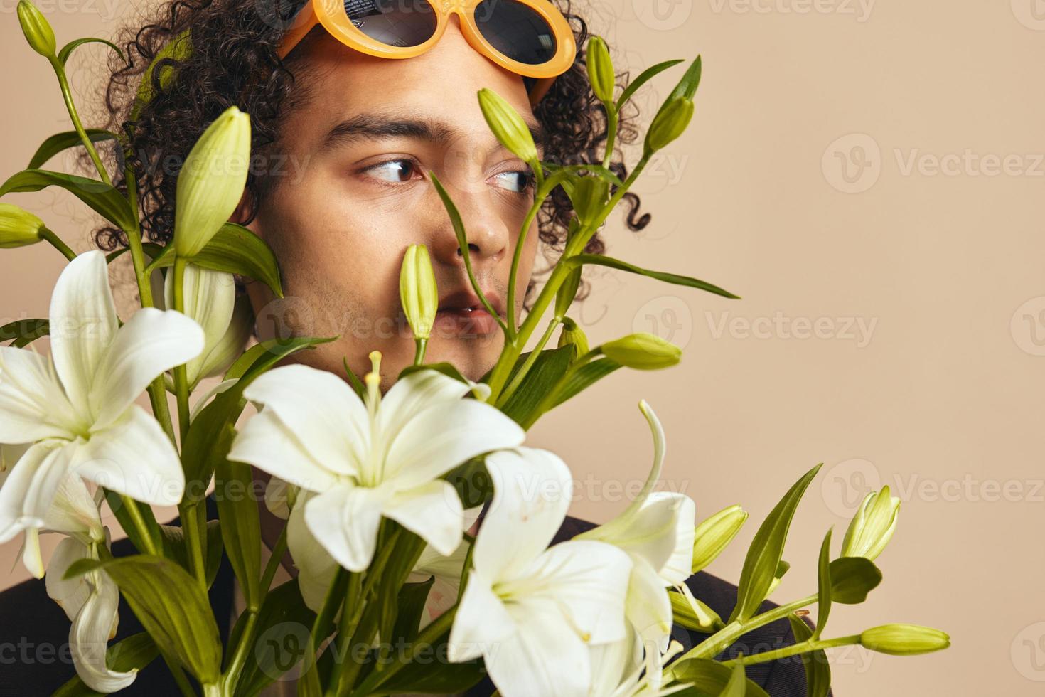 de cerca retrato de hermoso bronceado Rizado hombre en de moda Gafas de sol sostener lirios cerca cara posando aislado en terminado beige antecedentes. Moda nuevo colección oferta. retro estilo concepto. gratis sitio para anuncio foto