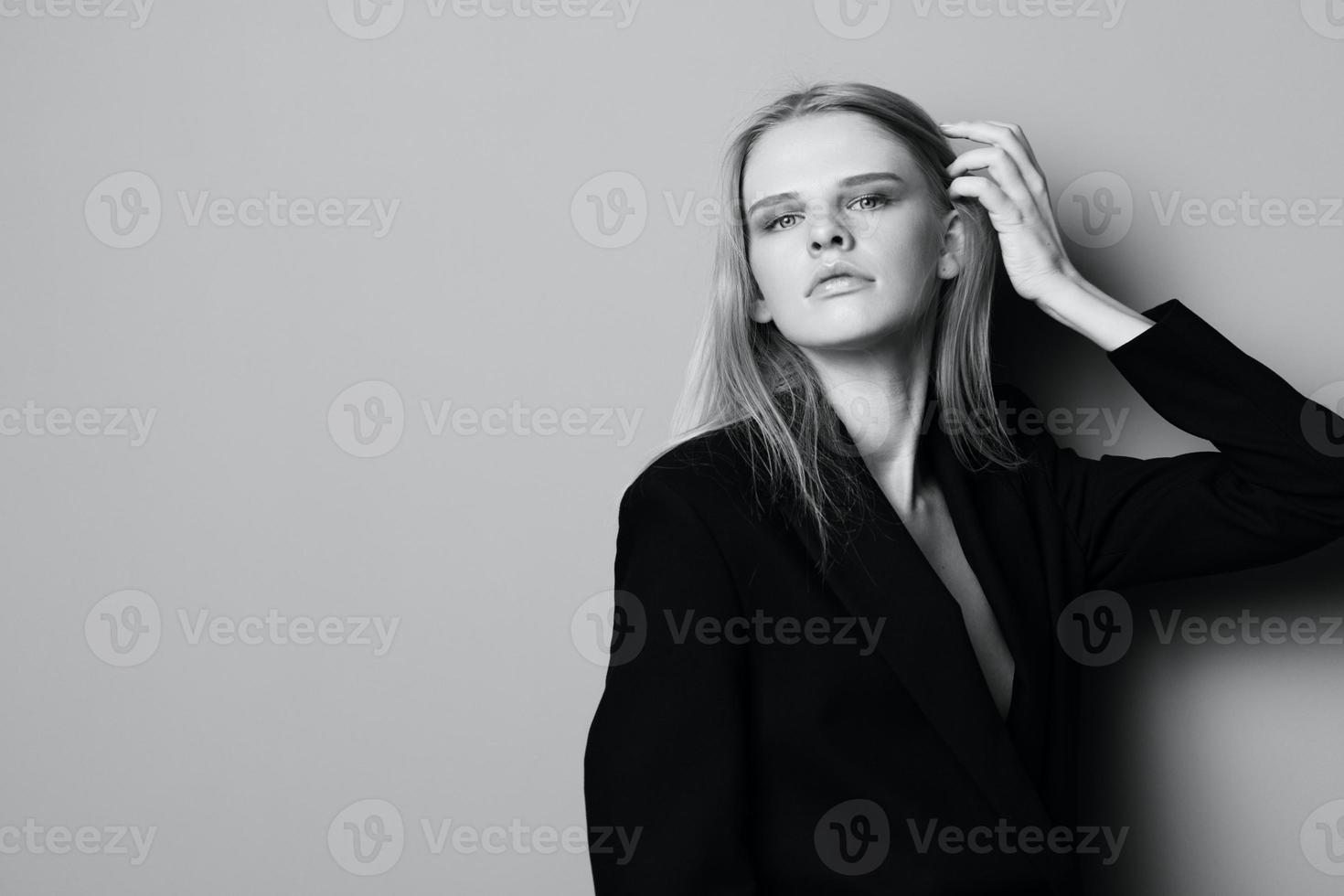 Sexy serious young woman in black jacket leans on the studio wall touching hair looks at camera. Stylish concept for fashion brands banner photo