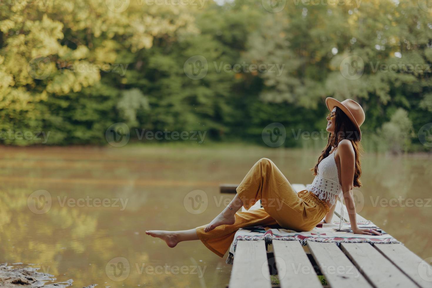 A hippie woman sits on a bridge by the river and enjoys the beautiful scenery around her cheerfully lifting her legs photo