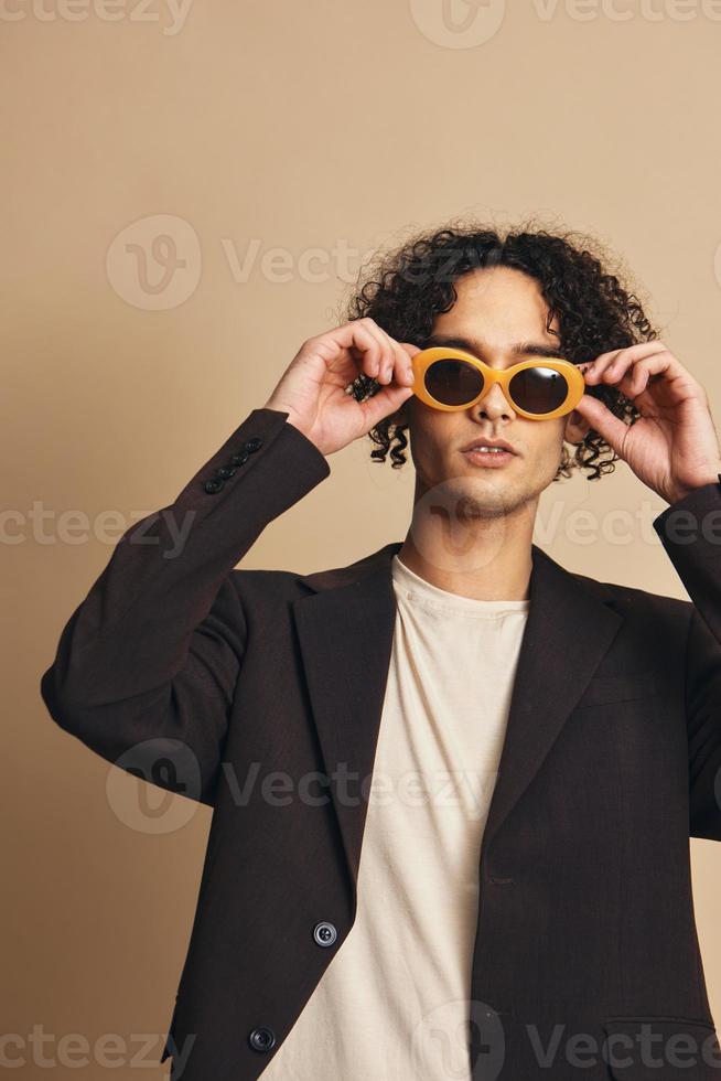 Demonstrative awesome tanned curly man in classic brown jacket trendy sunglasses posing isolated on over beige pastel background. Fashion New Collection offer. Retro style concept. Free place for adDemonstrative awesome tanned curly man in classic brown j photo