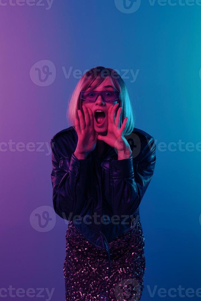 Excited sexy beautiful blonde woman in leather jacket sparkly dress trendy sunglasses open mouth touch cheek posing isolated in color light studio background. Neon party Cyberpunk concept. Copy space photo
