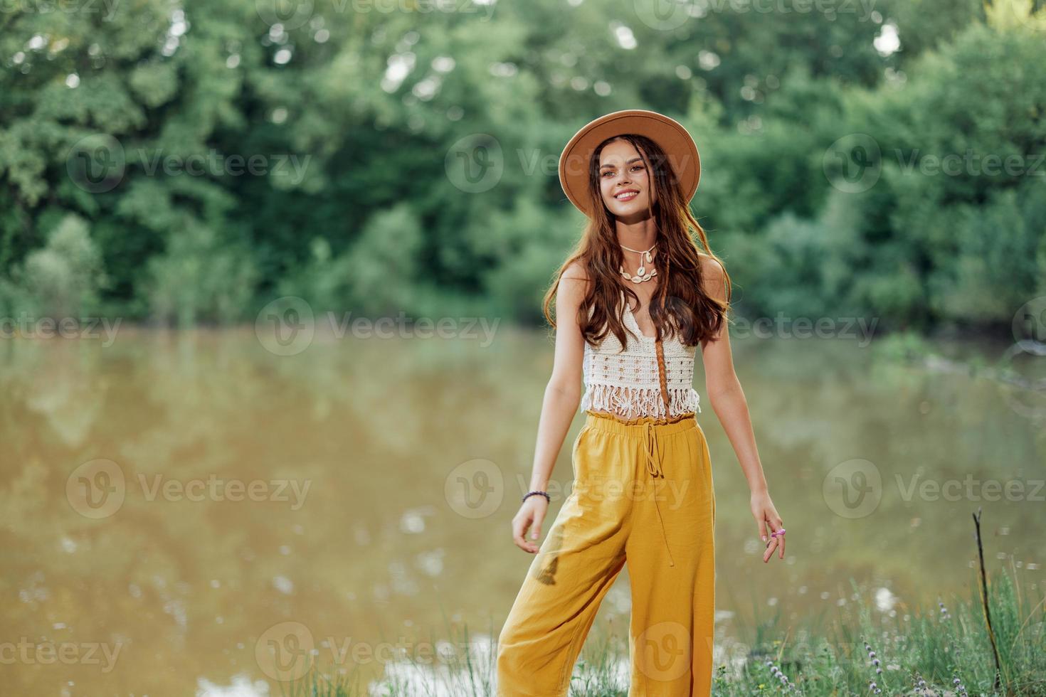 A young woman in a hippie look travels in nature by the lake wearing a hat and yellow pants in the fall photo