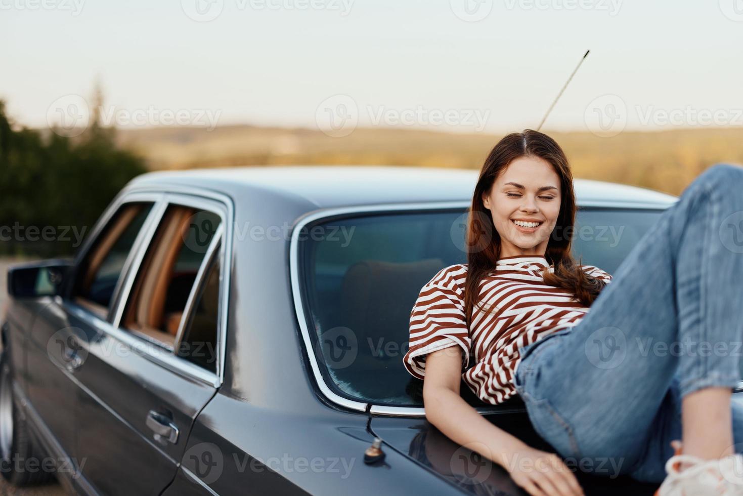 The woman driver stopped on the road and lay down on the car to rest and look at the beautiful landscape in a striped T-shirt and jeans. Complicated journey to nature photo