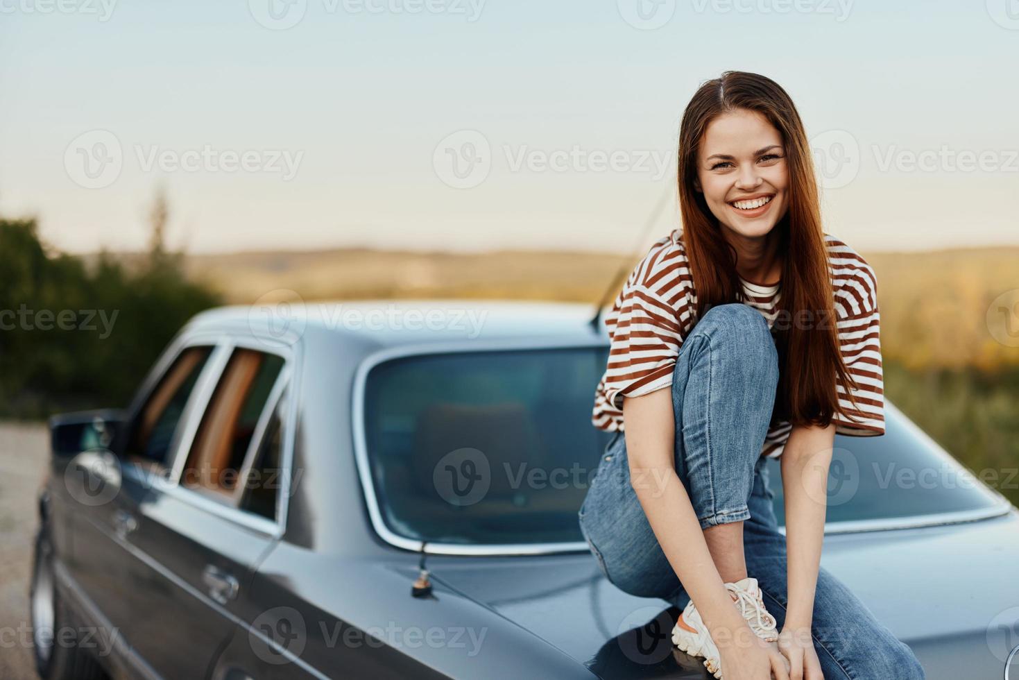 A young woman sits on the trunk of a car, laughs and rests after a difficult road and admires nature with a beautiful view. Stopping is also part of the journey photo
