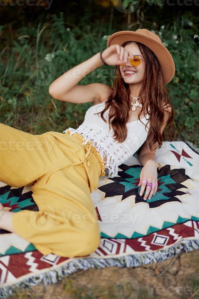 Hippie woman smiling in eco clothing yellow pants, white knit top, hat and yellow glasses sitting on plaid in park watching sunset, lifestyle camping trip photo