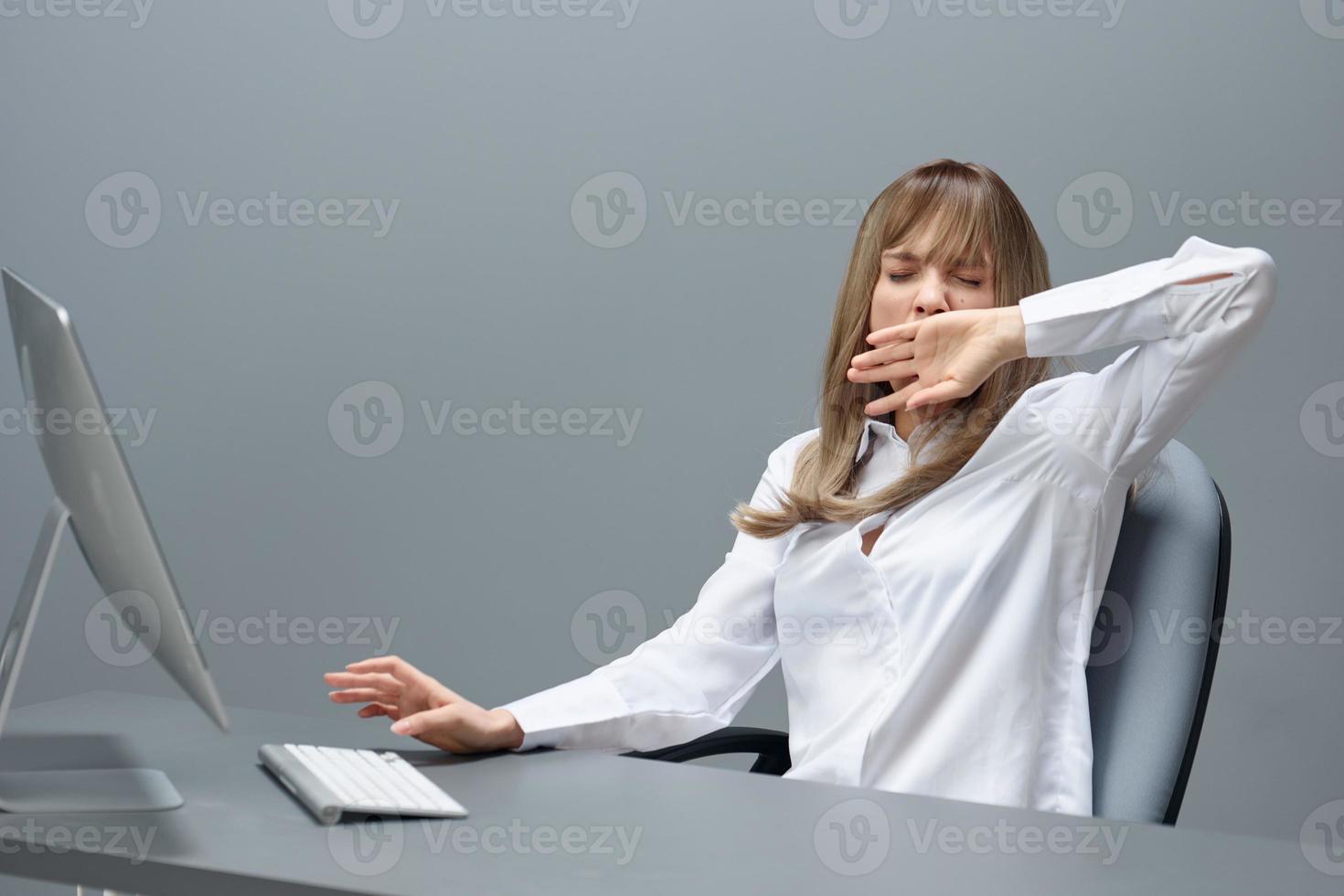 Sleepy tired pretty blonde businesswoman worker freelancer using desktop computer yawns sitting at workplace in gray modern office. Remote Job, Technology And Career Profession Concept. Copy space photo