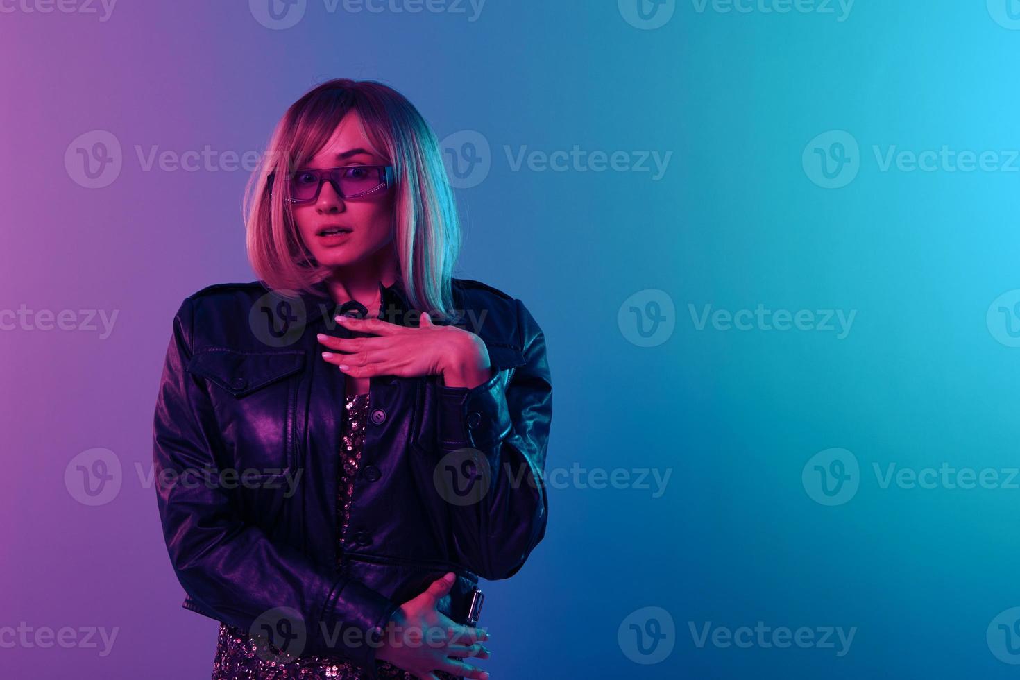 Shocked beautiful blonde woman in leather jacket sparkly dress trendy sunglasses hold hand on chest posing isolated in blue pink color light background. Neon party Cyberpunk concept. Copy space photo
