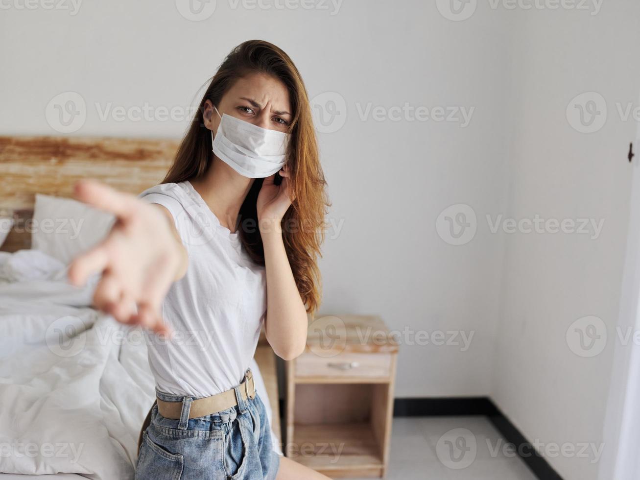 indignant woman with medical mask on her face sits on bed on vacation coronavirus quarantine photo