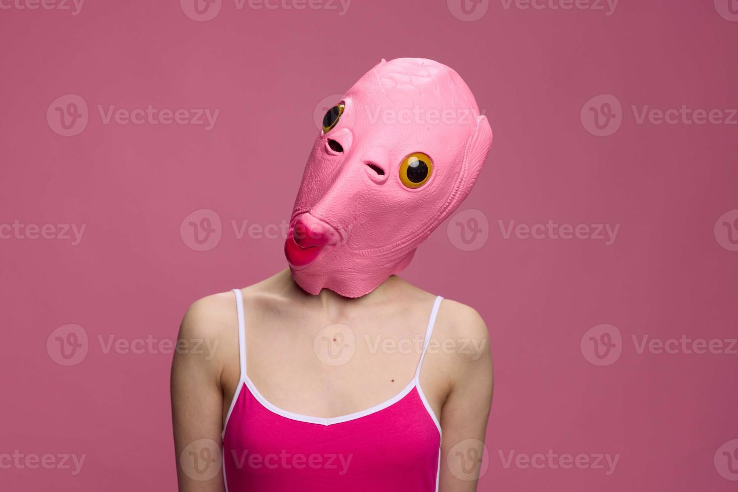 Woman wearing a pink fish head Halloween mask stands and looks at the camera against a pink background photo