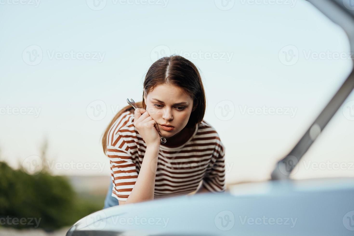 A woman with a wrench looks sadly and sadly under the open hood of her car and can't fix it from a roadside breakdown while traveling alone photo