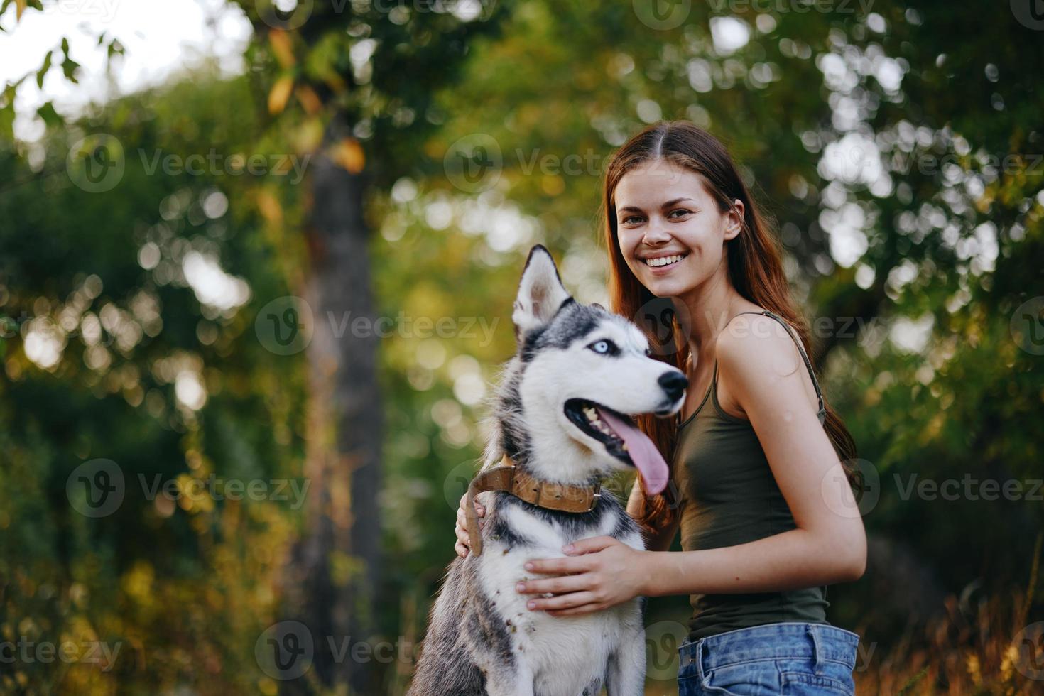 A woman with a husky breed dog smiles and affectionately strokes her beloved dog while walking in nature in the park in autumn against the backdrop of sunset photo