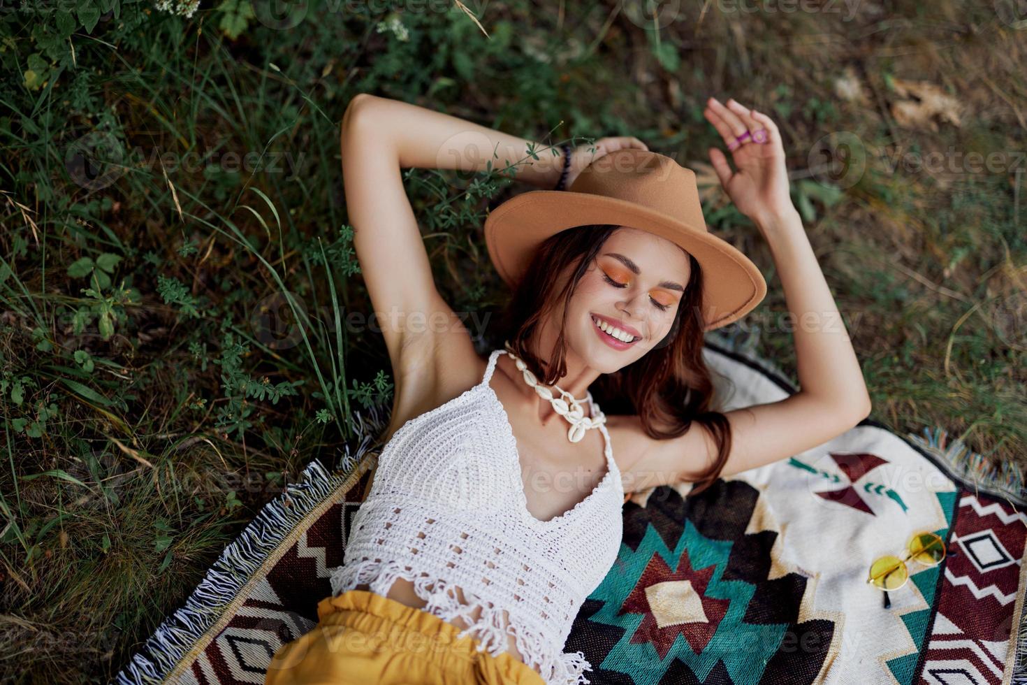 A woman in eco-friendly clothing in a hippie look is lying on a colored plaid smiling and looking at an autumn sunset in nature in the park. Lifestyle on a journey of harmony with the world photo