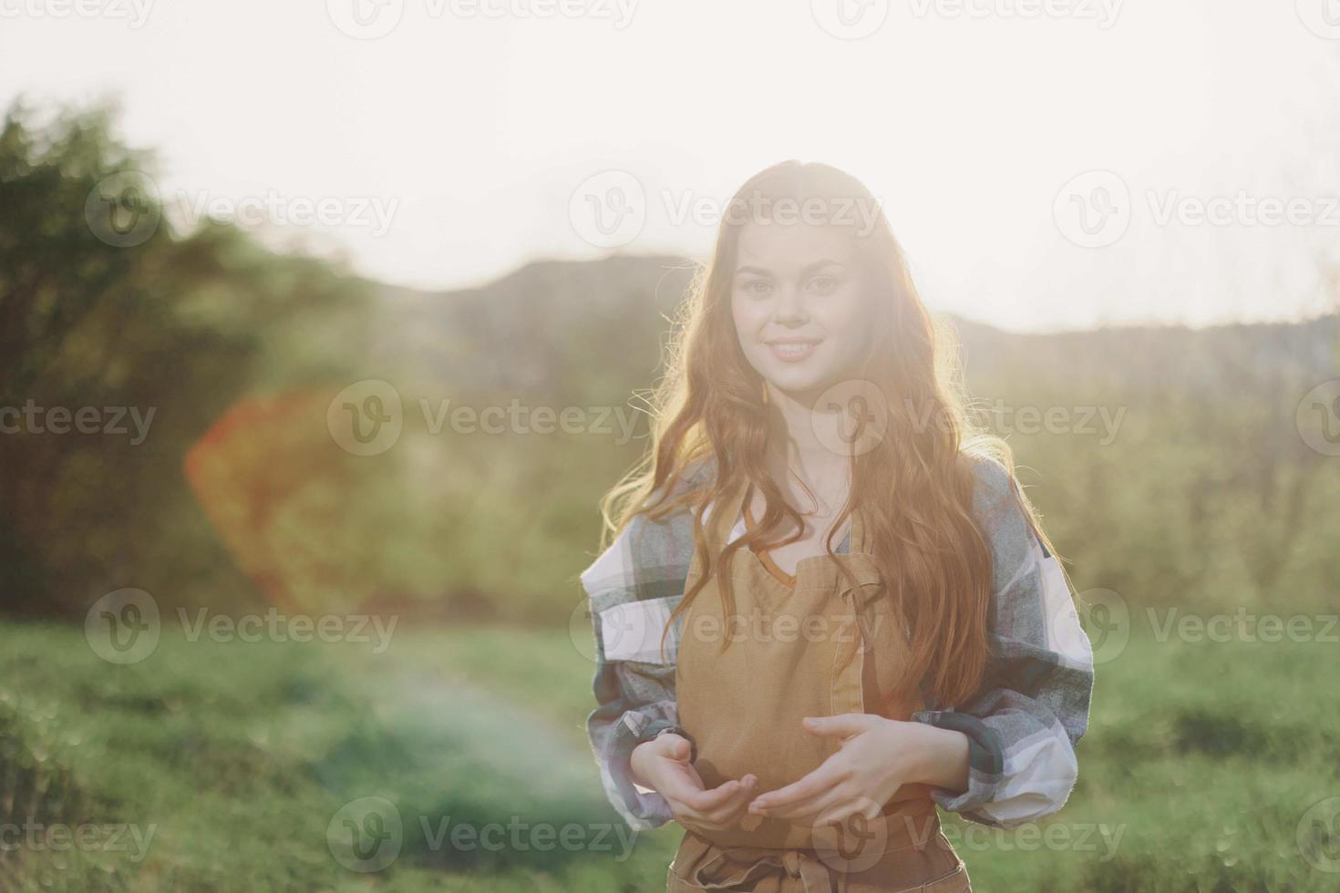 A woman gardener in an apron stands in a field of green grass outdoors, smiling on a summer afternoon into a sunny sunset after a day's work photo