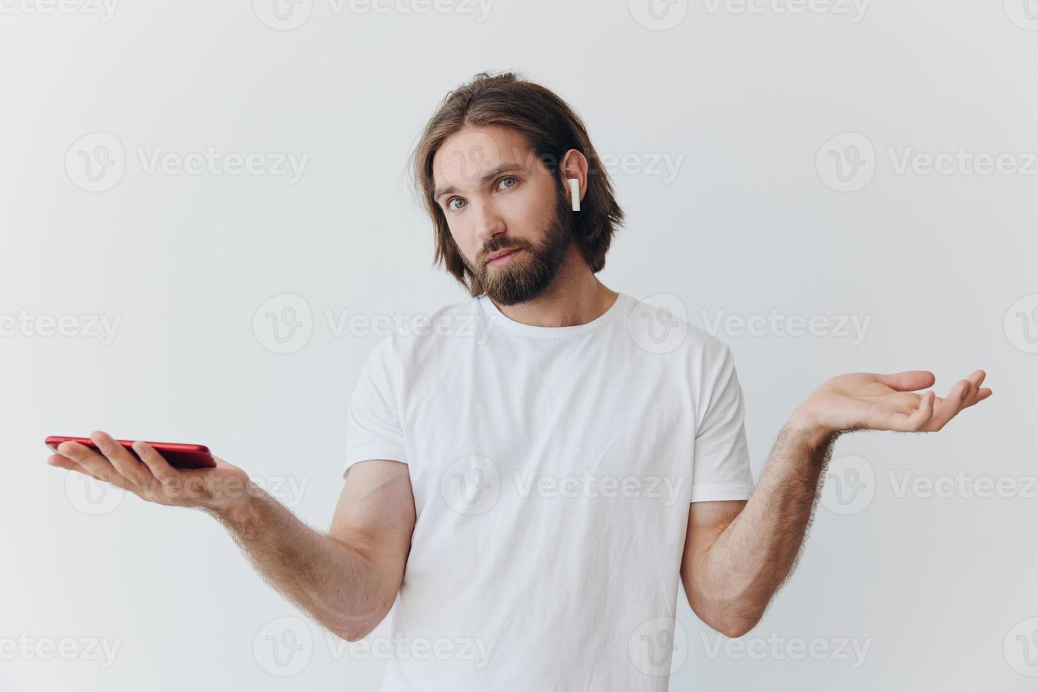 A man with a beard blogger in a white T-shirt with a phone and wireless headphones looks into the phone and spreads his hands to the side on a white background isolated copy space photo