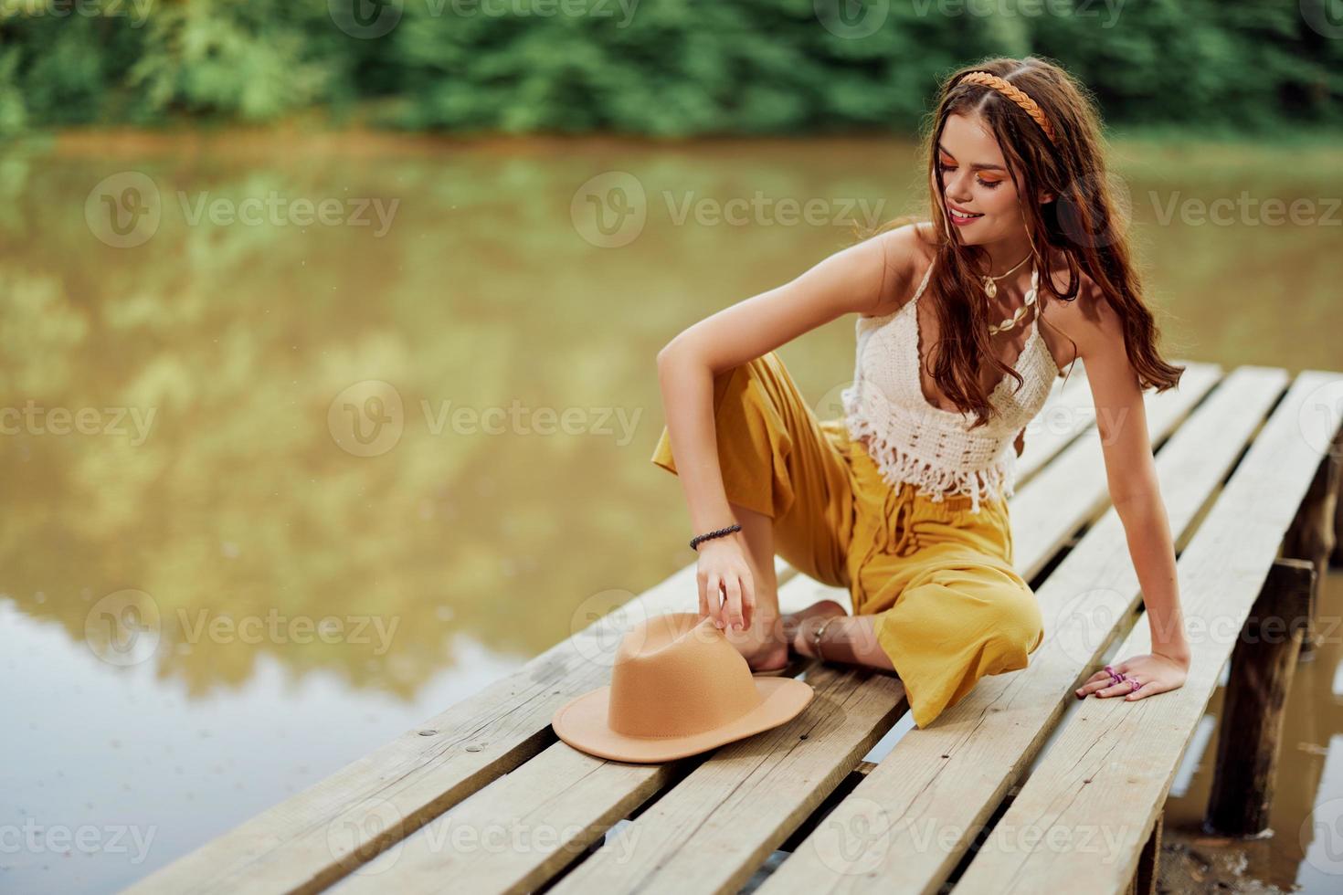 A young hippie woman sits on a lake bridge wearing stylish eco clothes and smiling photo