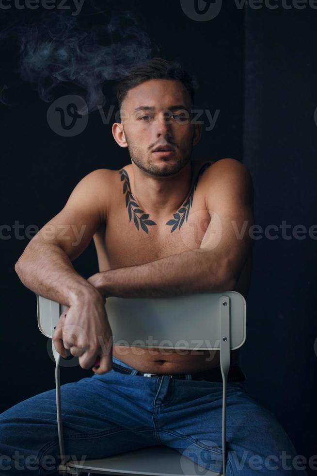 Modelling snapshots. Pensive frustrated tanned attractive handsome naked man sit on chair smoking looks at camera posing isolated in black studio background. Fashion offer. Copy space for ad. Closeup photo