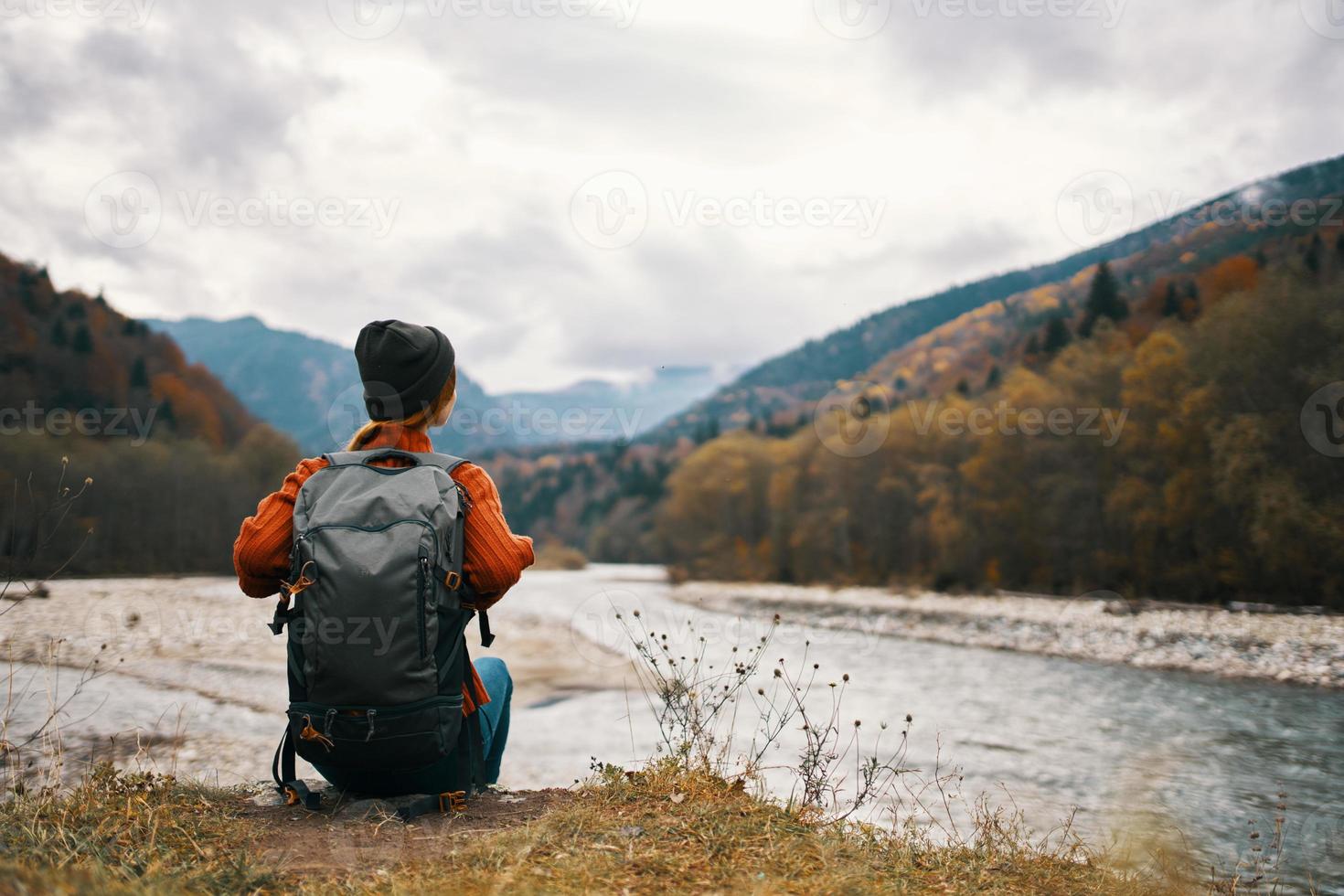 woman with a backpack and in a jacket with a hat on the river bank in the mountains in nature photo