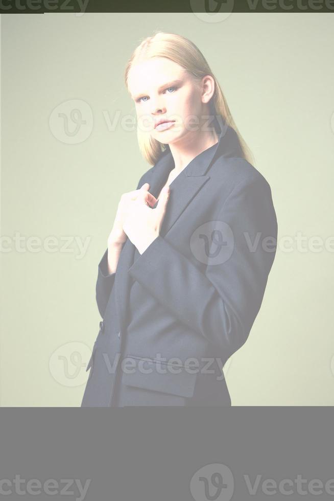 The blonde beauty holds her jacket by the collar half-turned posing on a green background in the studio. Concept photo for clothing brands. Cool offer for fashionable suits