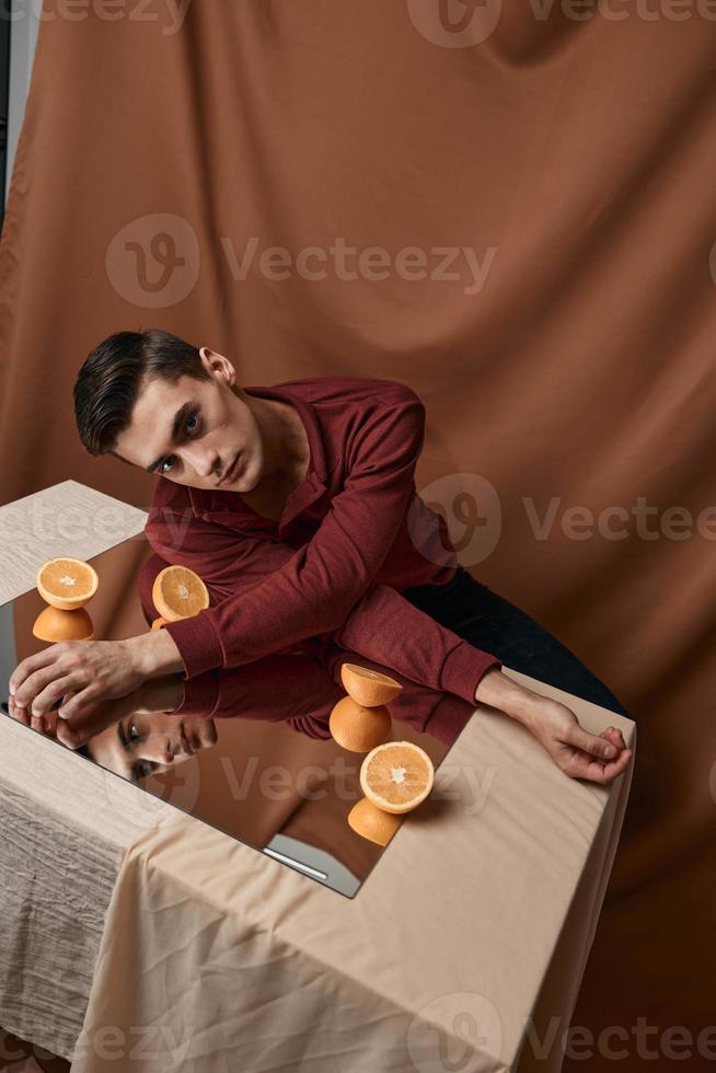 A guy in a shirt sits at a table with a mirror and orange oranges fabric background studio model photo