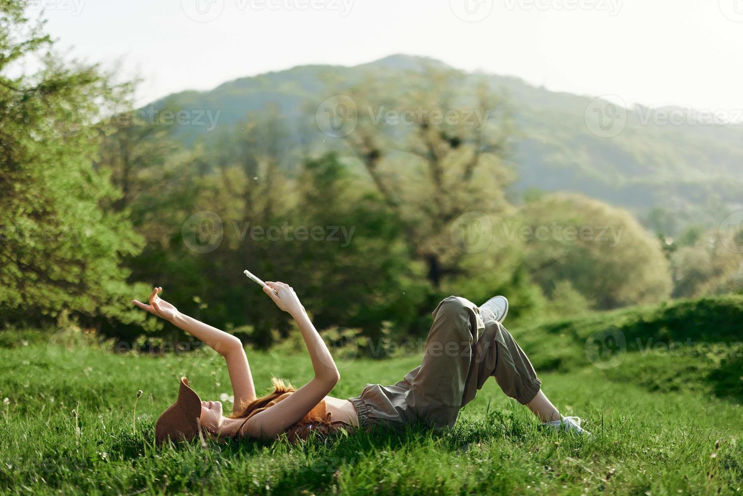 Happy woman blogger lying on the grass in the park and smiling with her phone in her hands against the backdrop of a summer natural landscape with sunlight photo