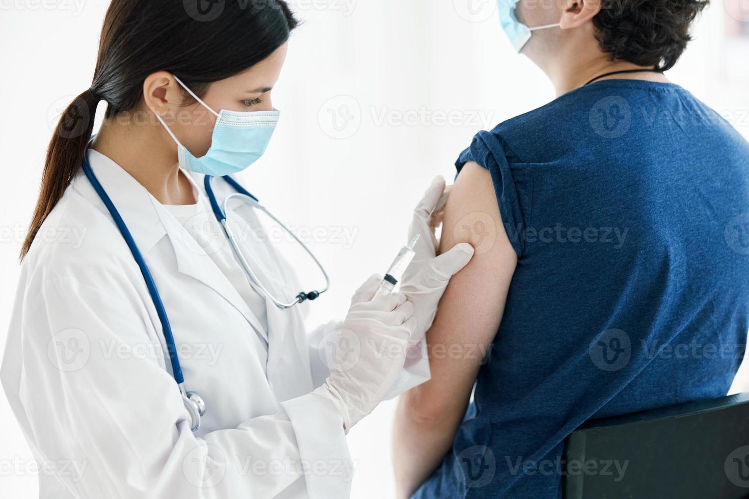 un enfermero inyecta un vacuna dentro el hombro de un paciente vistiendo un médico máscara foto