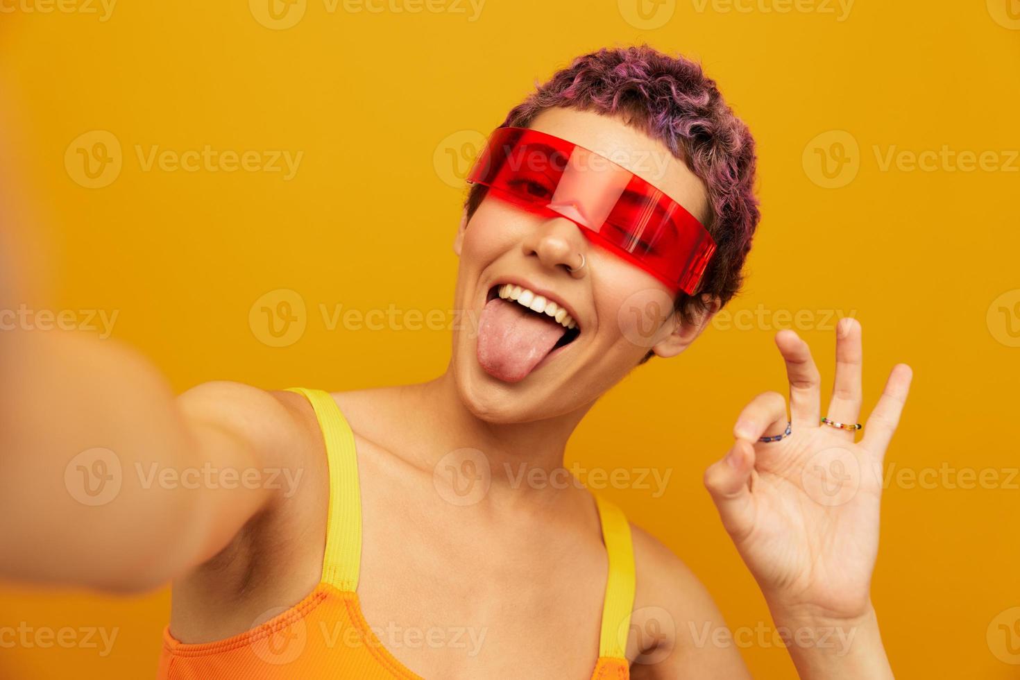 Woman wearing unusual millennial glasses taking selfies in sportswear against an orange studio background, free space photo