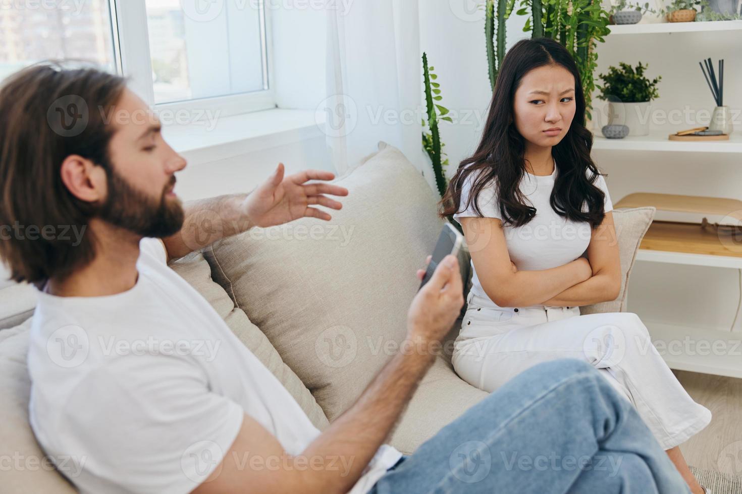 un hombre mira a el teléfono pantalla durante un argumento con su novia. el enojado y herir mujer mira en su dirección y es triste. familia discordia a hogar, teléfono adiccion foto