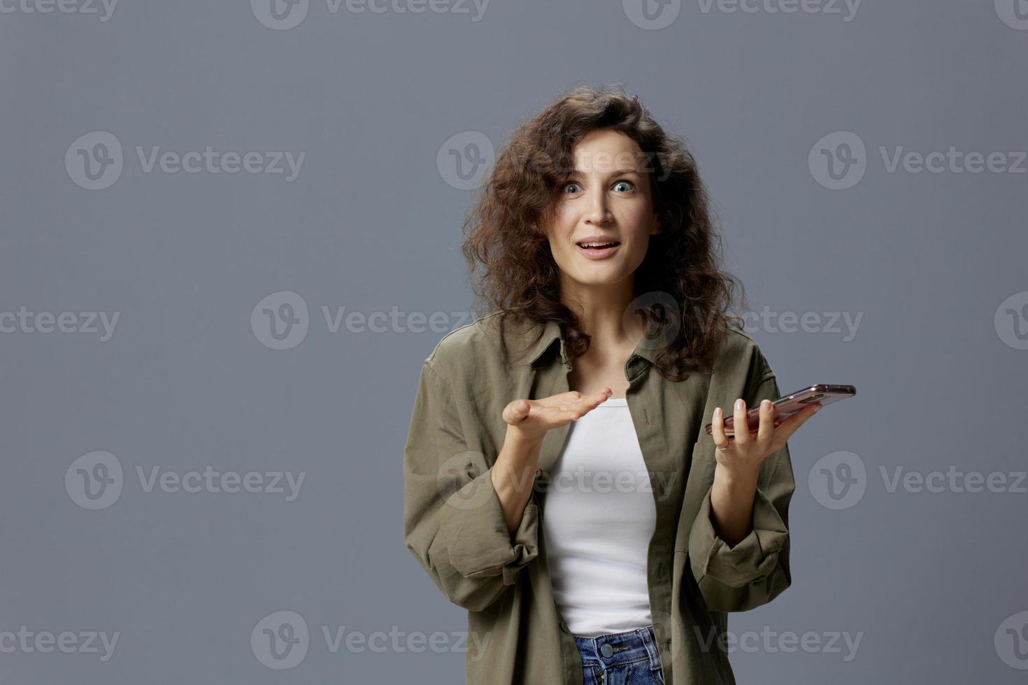 emocionado contento Rizado hermosa mujer en casual caqui verde camisa conmocionado desde Noticias ese leer utilizando teléfono posando aislado en terminado gris azul antecedentes. social medios de comunicación hombre de influencia concepto. Copiar espacio foto