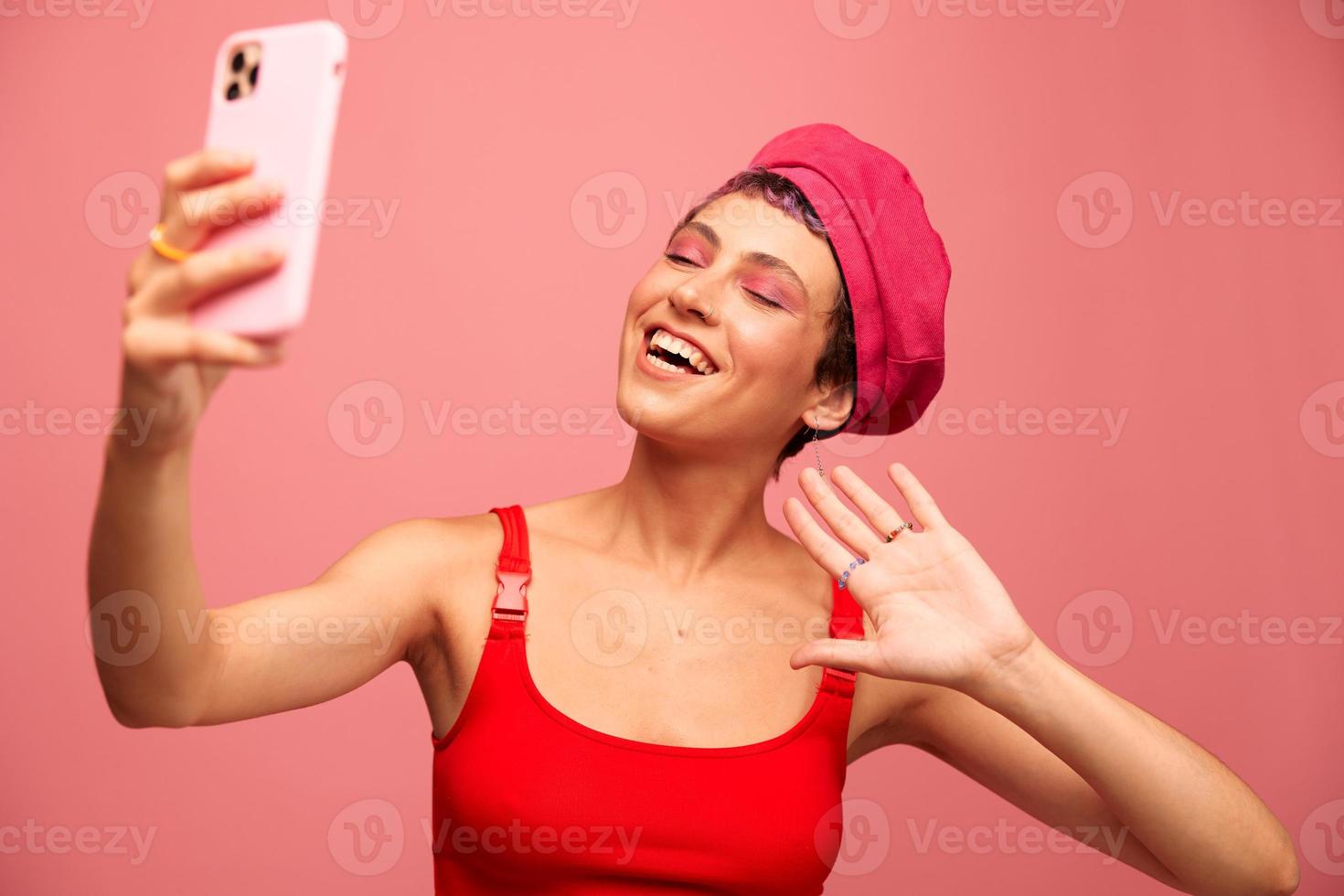 A young woman blogger with colored pink hair and a short haircut takes a picture of herself on the phone and broadcasts a smile in stylish clothes and a hat on a pink background monochrome style photo