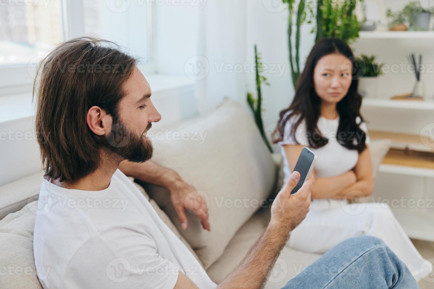 un hombre mira a su teléfono pantalla y sonrisas durante un argumento con su novia. el enojado y herir mujer mira en su dirección y es triste. familia discordia a hogar, teléfono adiccion foto