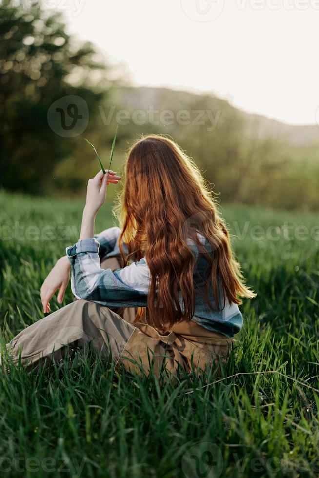 un mujer se sienta en el verde césped en un parque con su espalda a el cámara y relaja en naturaleza en el verano puesta de sol noche ligero foto