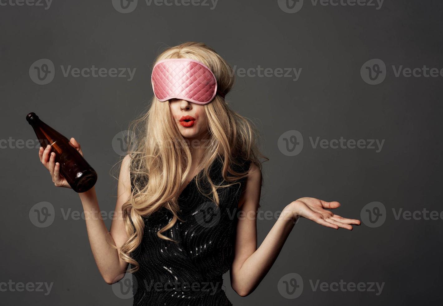 Drunk woman with a bottle of alcohol in her hand on a gray background and a mask for sleeping photo