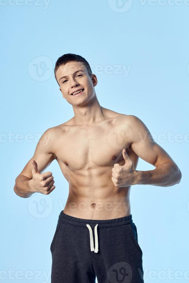 a guy with a naked torso and in shorts shows his thumb on a blue background photo