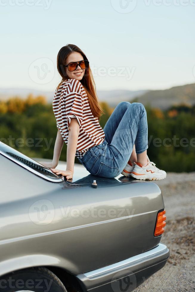 A fashion woman in stylish glasses, a striped t-shirt and jeans sits on the trunk of a car and looks at the beautiful nature of autumn photo