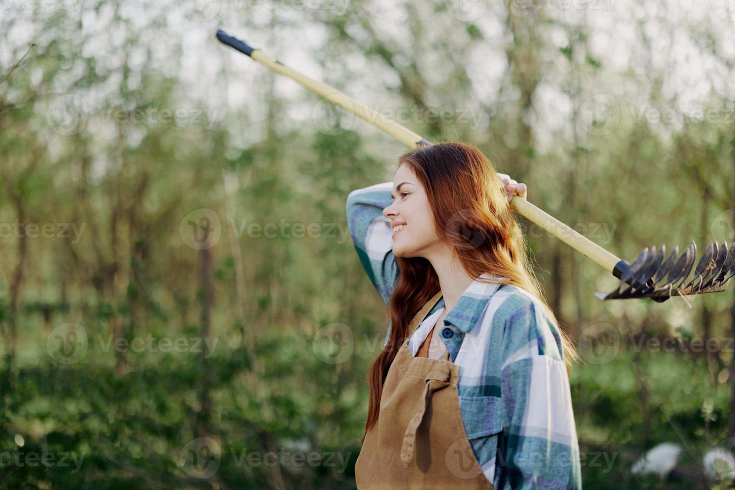 A woman smiling beautifully and looking at the camera, a farmer in work clothes and an apron working outdoors in nature and holding a rake to gather grass and forage for the animals in the garden photo