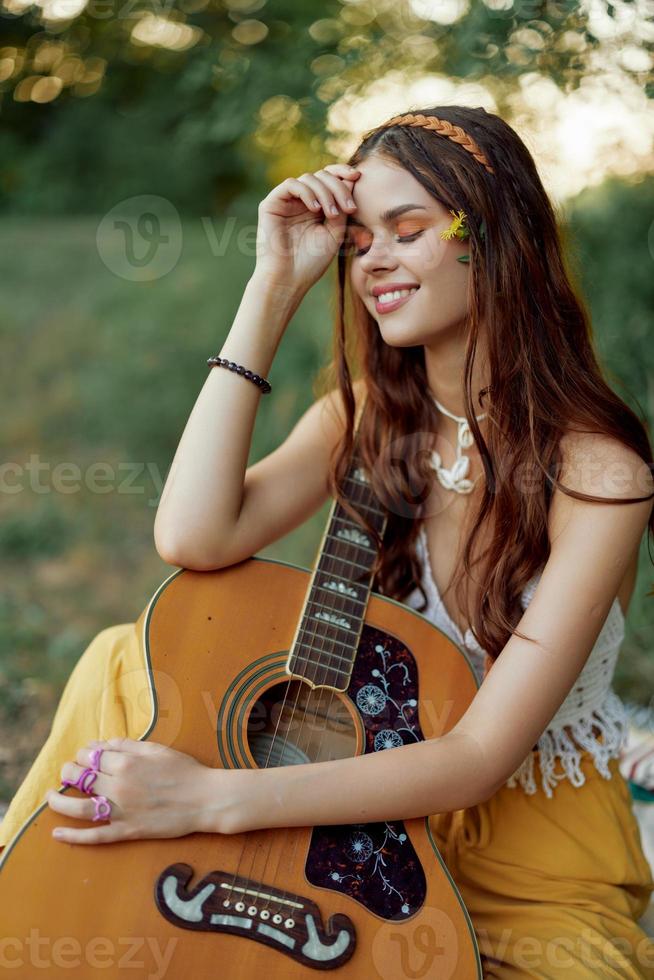 Hippie eco friendly woman with a guitar relaxing in nature sitting on a plaid by the lake in the evening in the rays of the setting sun. A lifestyle in harmony with the body and nature photo