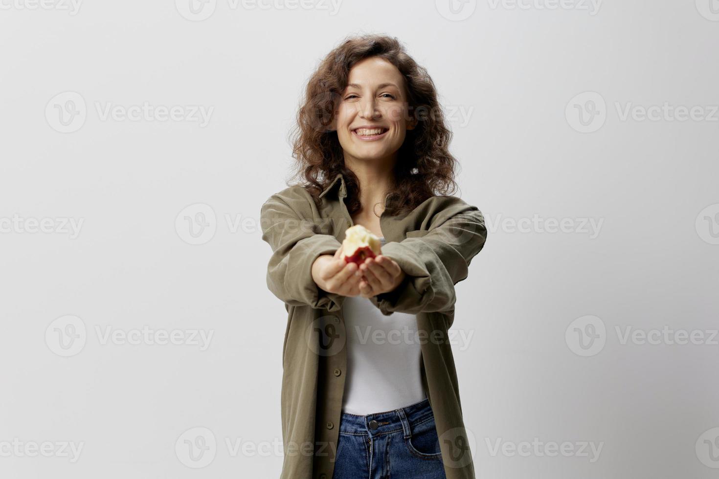 Delish it. Happy young curly beautiful woman in casual khaki green shirt shares apple, pulls it at camera posing isolated on over white background. Natural Eco-friendly products concept. Copy space photo