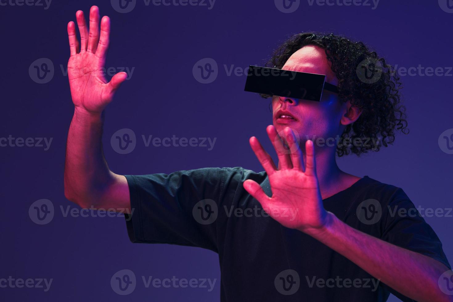 emocionado elegante bronceado Rizado hombre en negro camiseta gafas toques invisible objeto posando aislado en color rosado azul antecedentes. frio Moda oferta. virtual realidad nuevo colección concepto. Copiar espacio foto