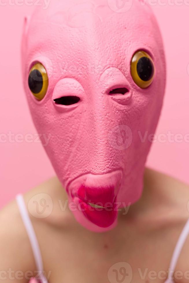 A woman wearing a silicone Halloween mask in the shape of a pink fish with big yellow eyes looks at the camera against a pink background photo