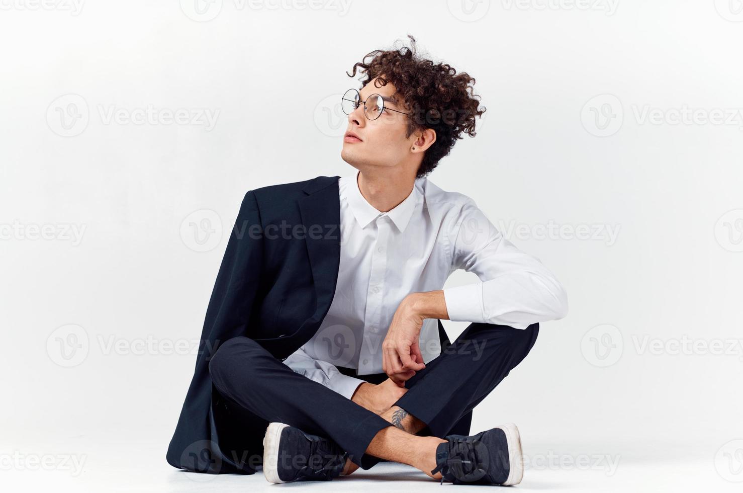 curly-haired guy in fashionable clothes and sneakers sits on the floor in a bright room in full growth photo
