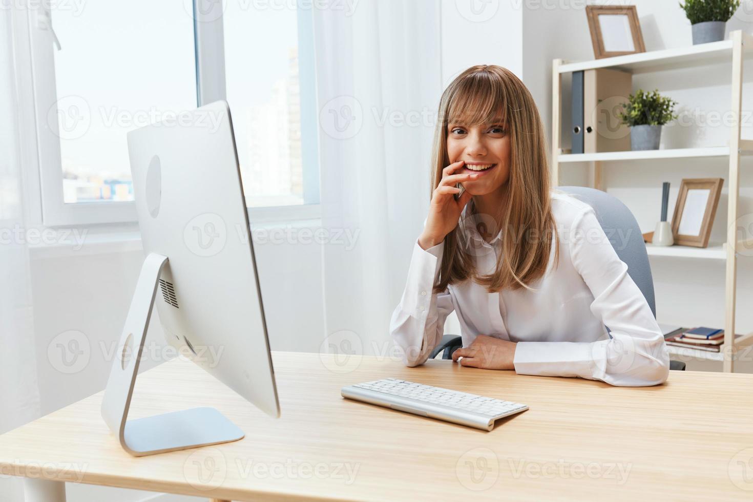 Happy smiling adorable blonde businesswoman worker freelancer look at camera sitting at workplace in light modern office. Cheerful employee work on computer online in support service. Copy space photo