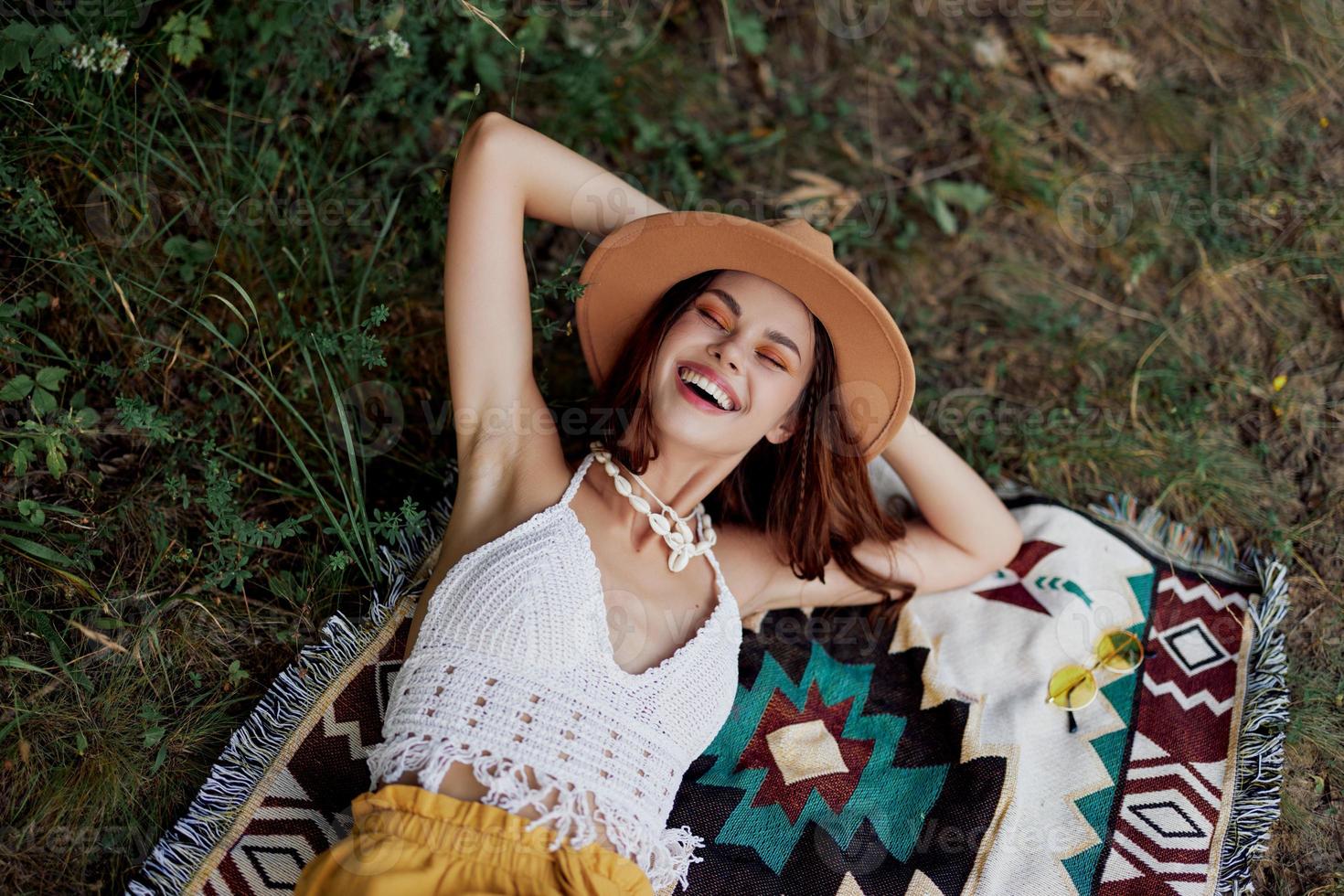 A woman in eco-friendly clothing in a hippie look is lying on a colored plaid smiling and looking at an autumn sunset in nature in the park. Lifestyle on a journey of harmony with the world photo