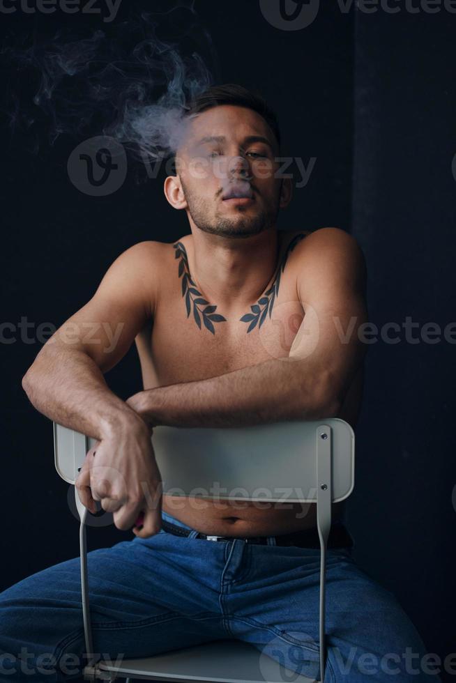 Modelling snapshots. Pensive serious tanned attractive handsome naked man sit on chair smoking looks aside posing isolated in black studio background. Fashion offer. Copy space for ad. Closeup photo