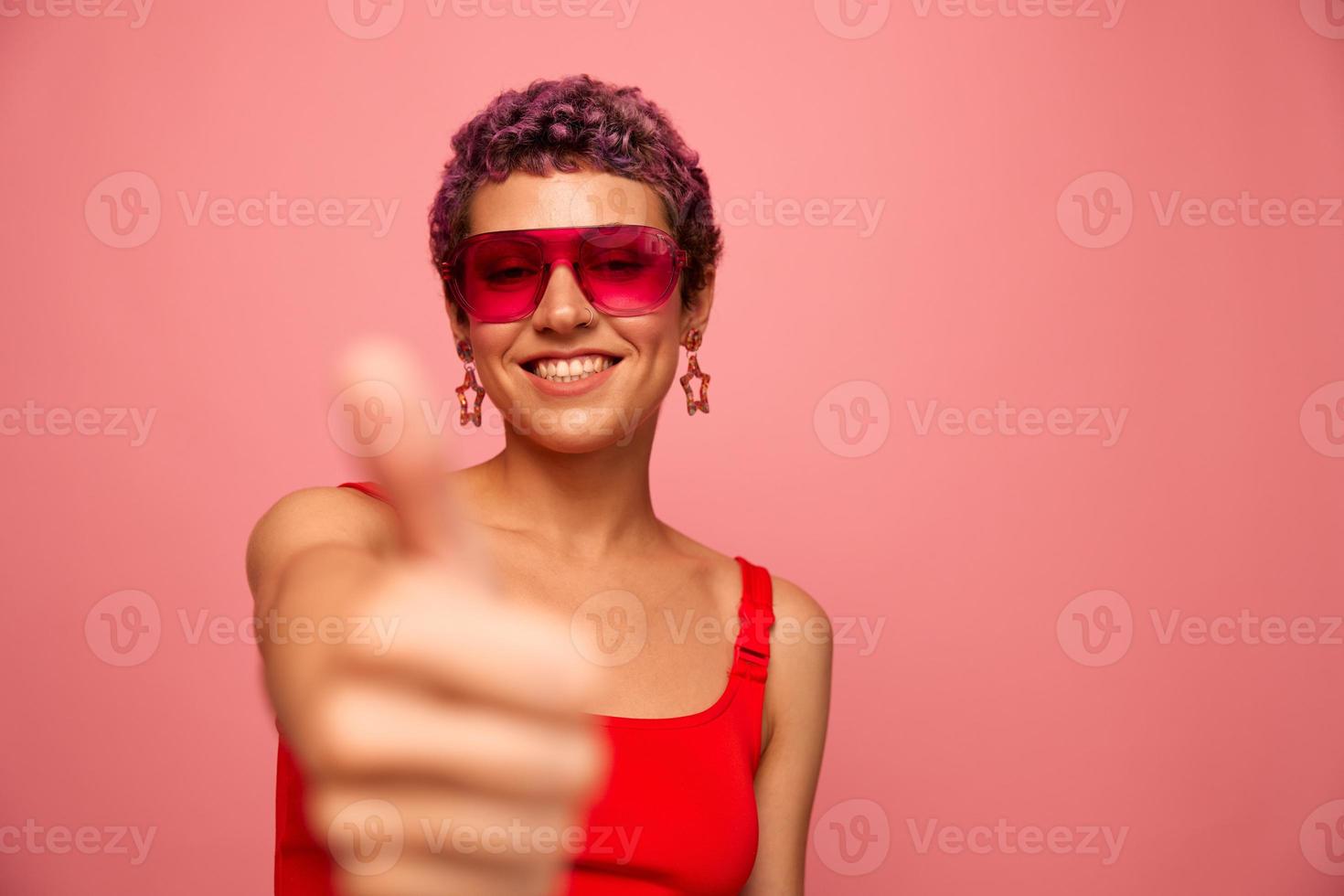 Fashion portrait of a woman with a short haircut in colored sunglasses with unusual accessories with earrings smiling on a pink bright background showing thumbs up at the camera photo