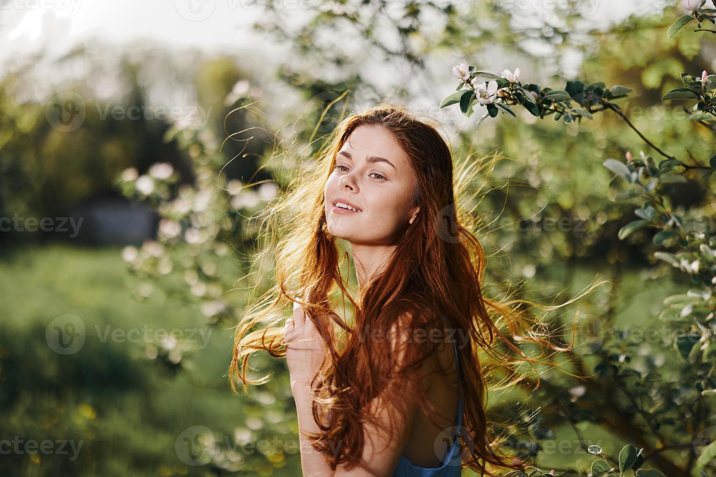 mujer con un hermosa sonrisa con dientes y largo pelo volador pelo en el primavera puesta de sol en naturaleza en el parque cerca el floración arboles felicidad, natural belleza y pelo salud foto