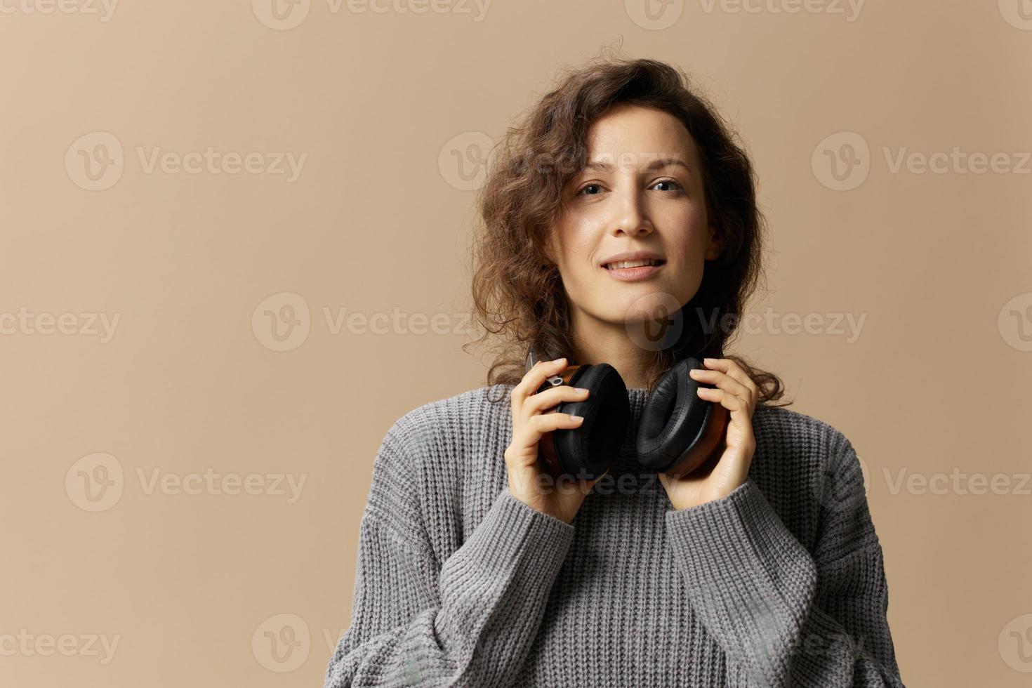 Cheerful enjoyed pretty curly beautiful female in gray casual sweater wear headphones looks at camera posing isolated on over beige pastel background. Sound streaming platform concept. Copy space photo