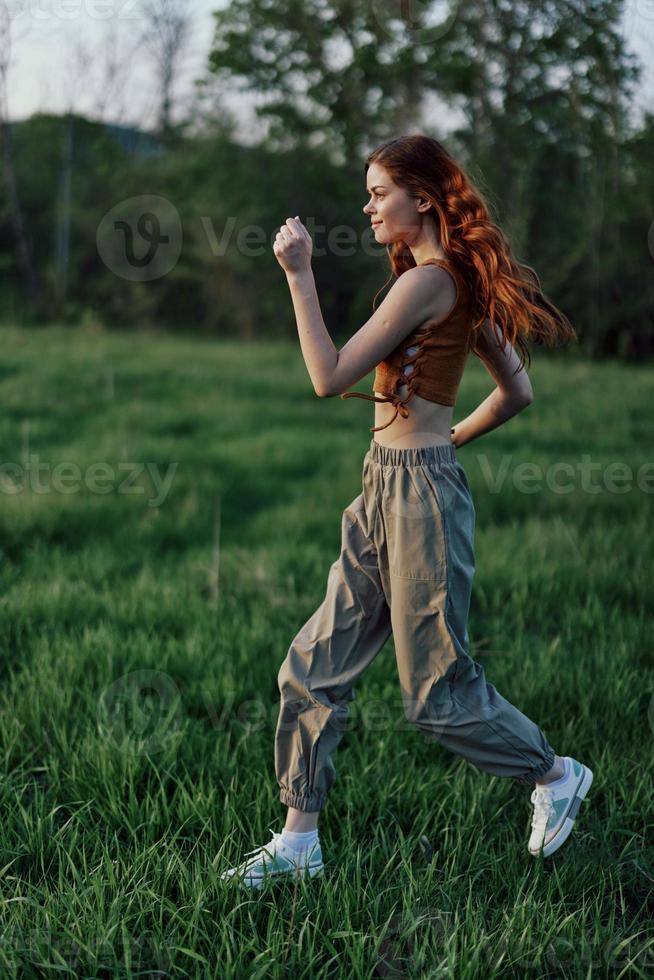 un mujer con largo rojo pelo trabajos fuera y carreras en el verde césped en el parque en pantalones deportivos y zapatillas foto