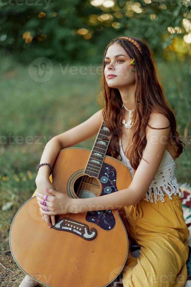 Hippie woman eco friendly playing guitar smiles and singing songs in nature sitting on a plaid by the lake in the evening in the sunset sunlight. A lifestyle in harmony with the body and nature photo