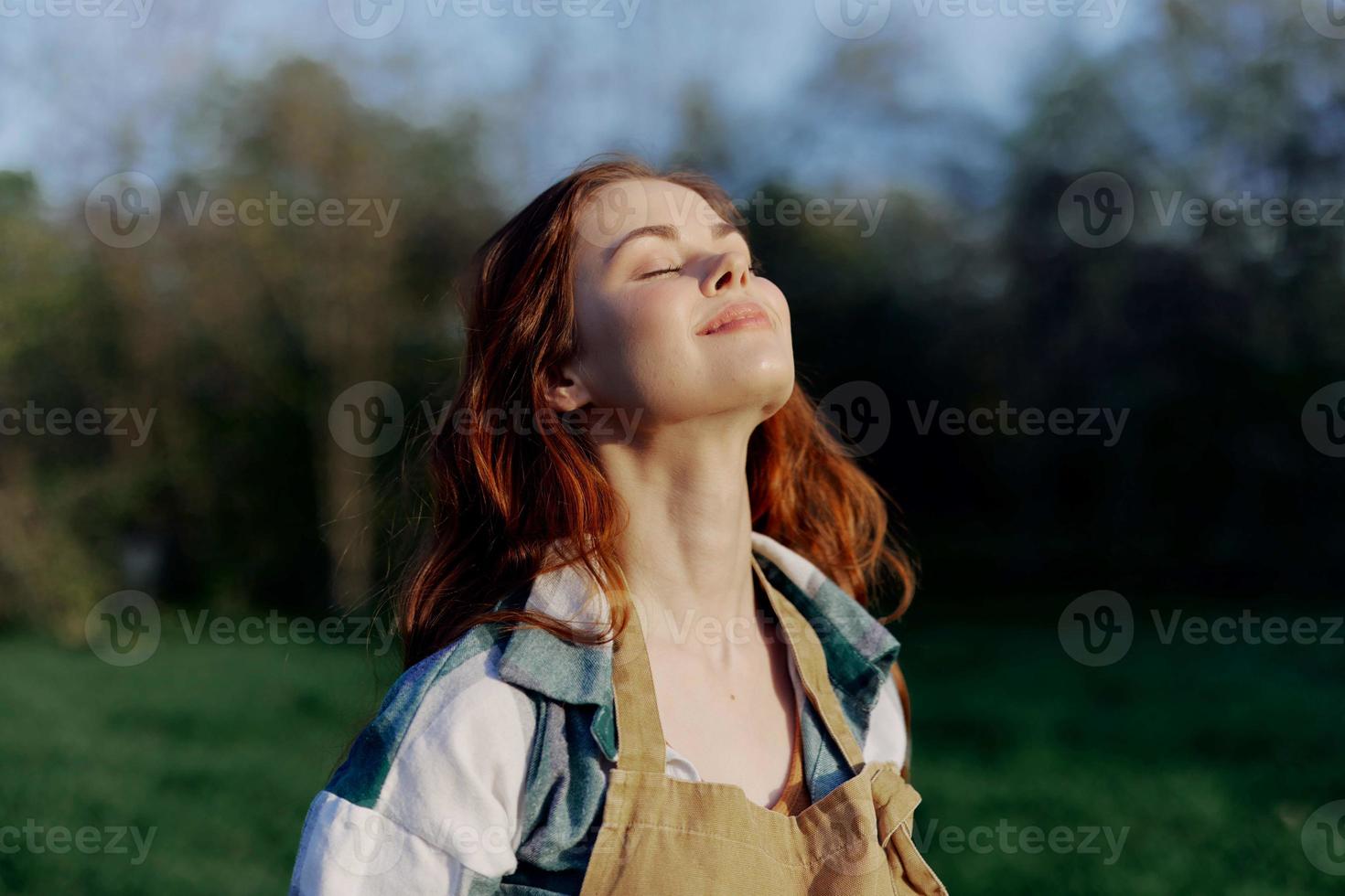 Portrait of a beautiful young woman with red hair in nature in a park in the summer, looking at the sunset against a background of green grass and trees photo