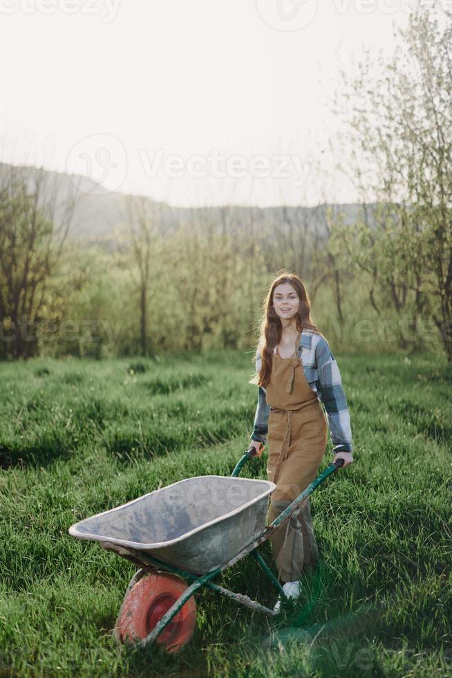 un contento mujer con un carro trabajos en su país hogar en el campo en contra un fondo de verde césped y puesta de sol Dom foto